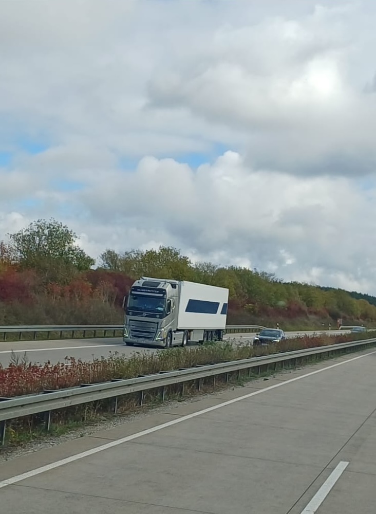Camion argintiu cu remorcă albă circulând pe o autostradă aglomerată.
